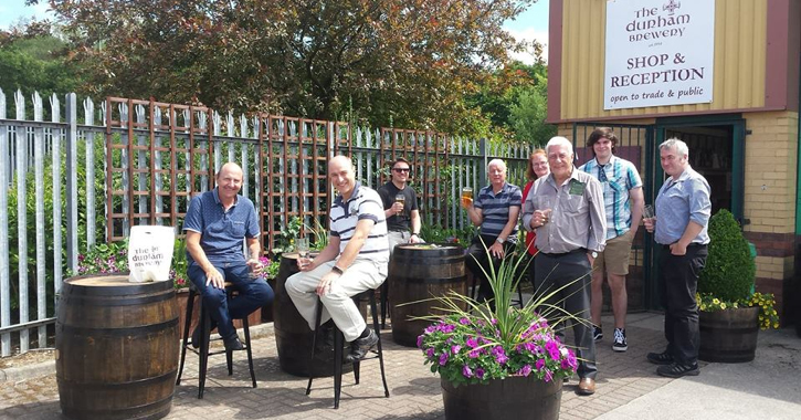 group of people sat outside Drurham brewery drinking beer and smiling at camera